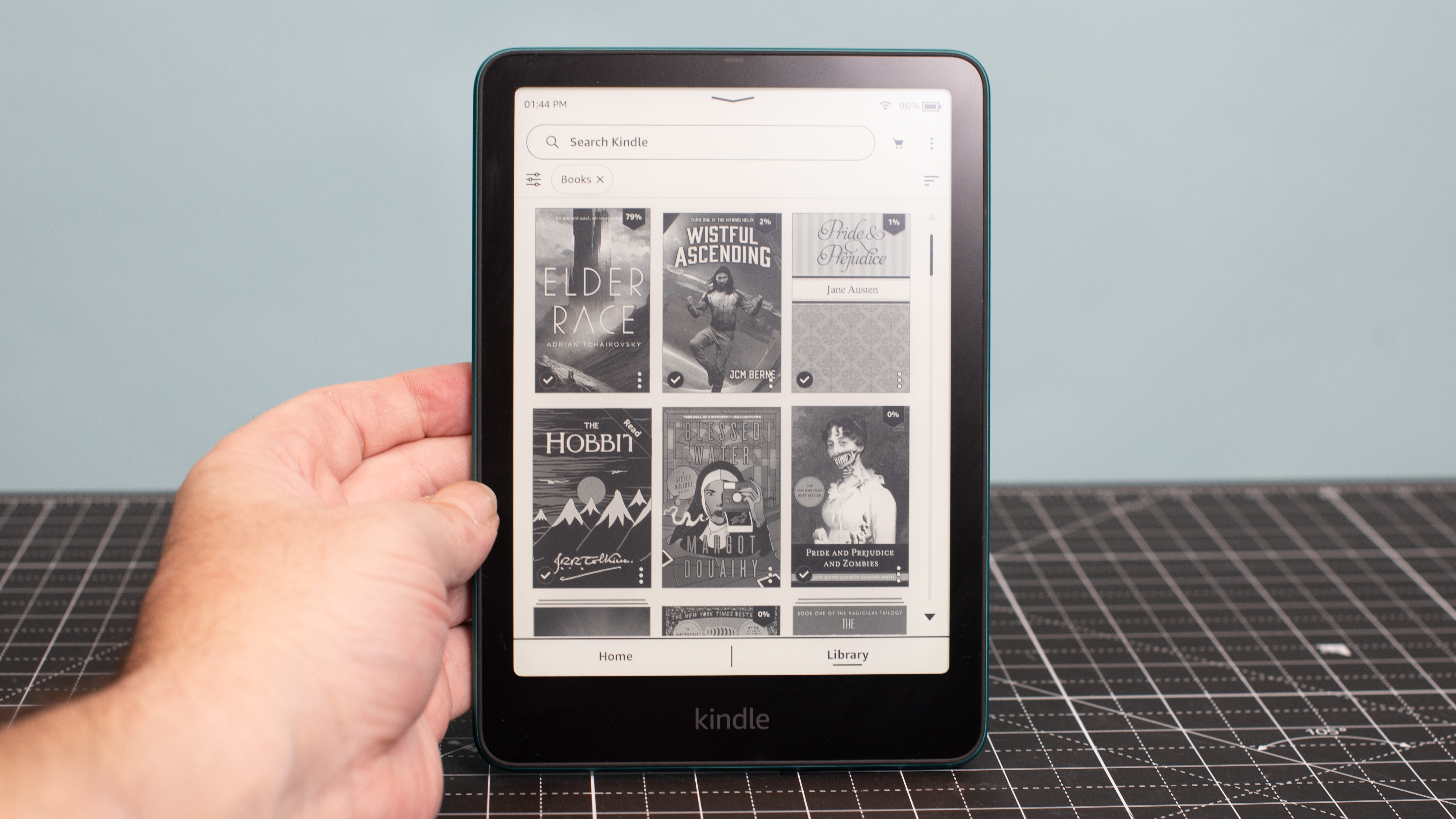 Kindle Paperwhite Signature Edition held up by one hand showing book covers in a grid including Elder Race by Adrian Tchaikovsky and Wistful Ascending by JCM Berne and The Hobbit by J.R.R. Tolkien