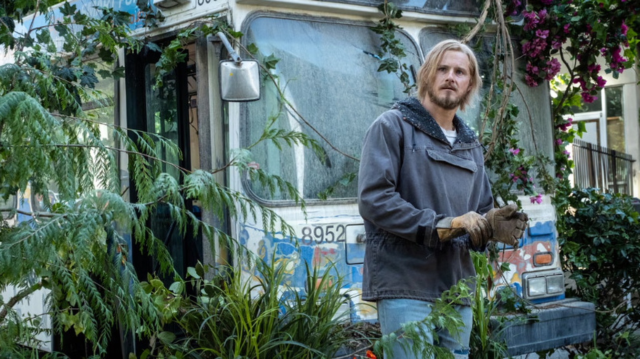 A disheveled looking Ish (Alexander Ludwig) stands in front of a beaten up bus in an apocalyptic city setting in Earth Abides