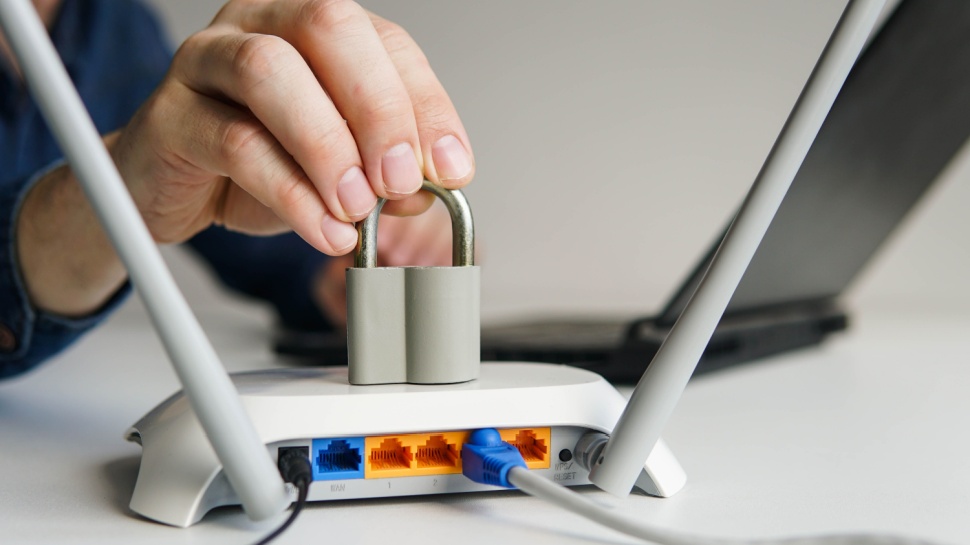 A Wi-Fi router placed on a desk with cables going in. A hand is holding a padlock on top of the router.