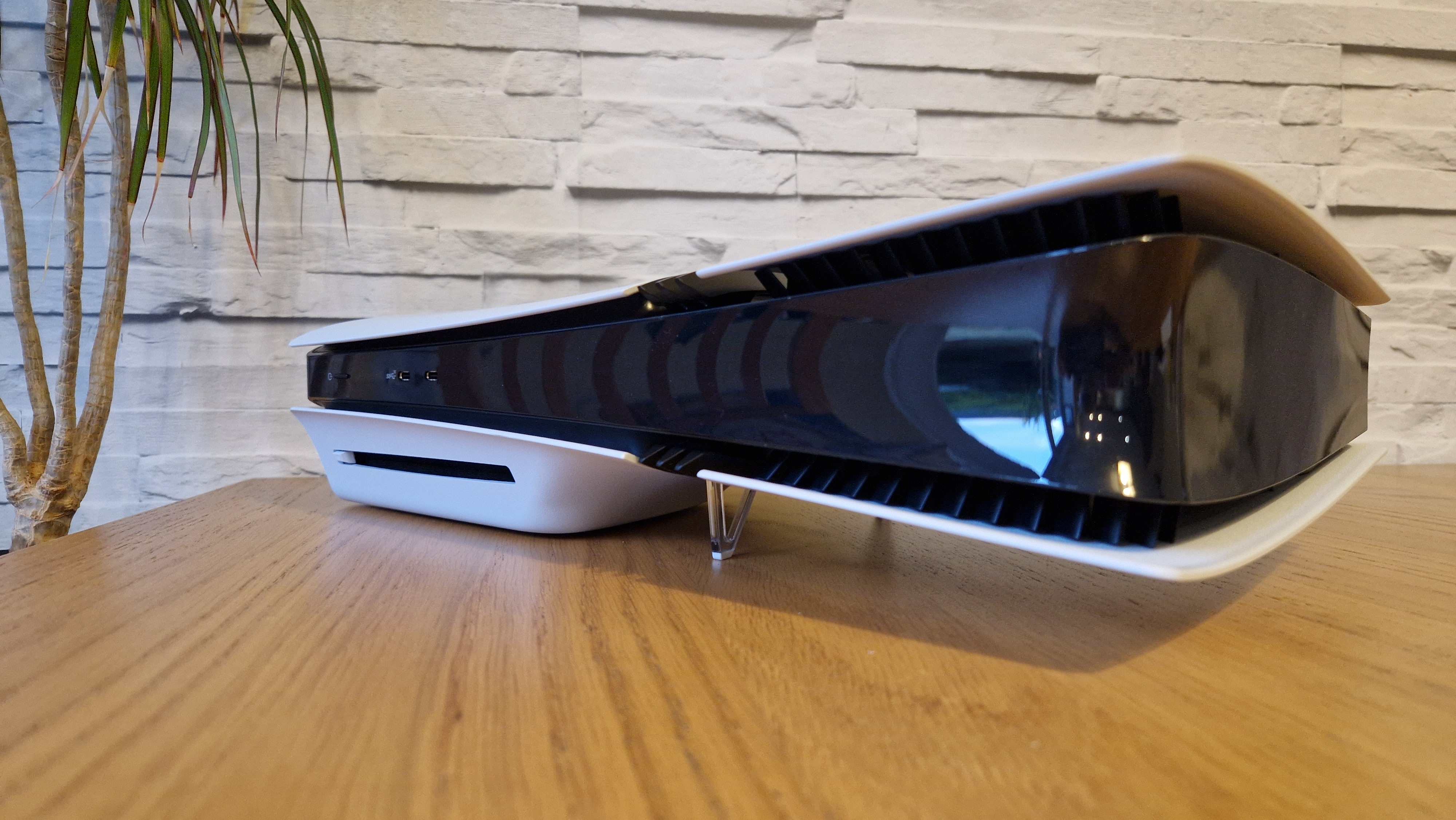 The PS5 Pro console on a wooden desk in front of a white brick background and next to a potted plant