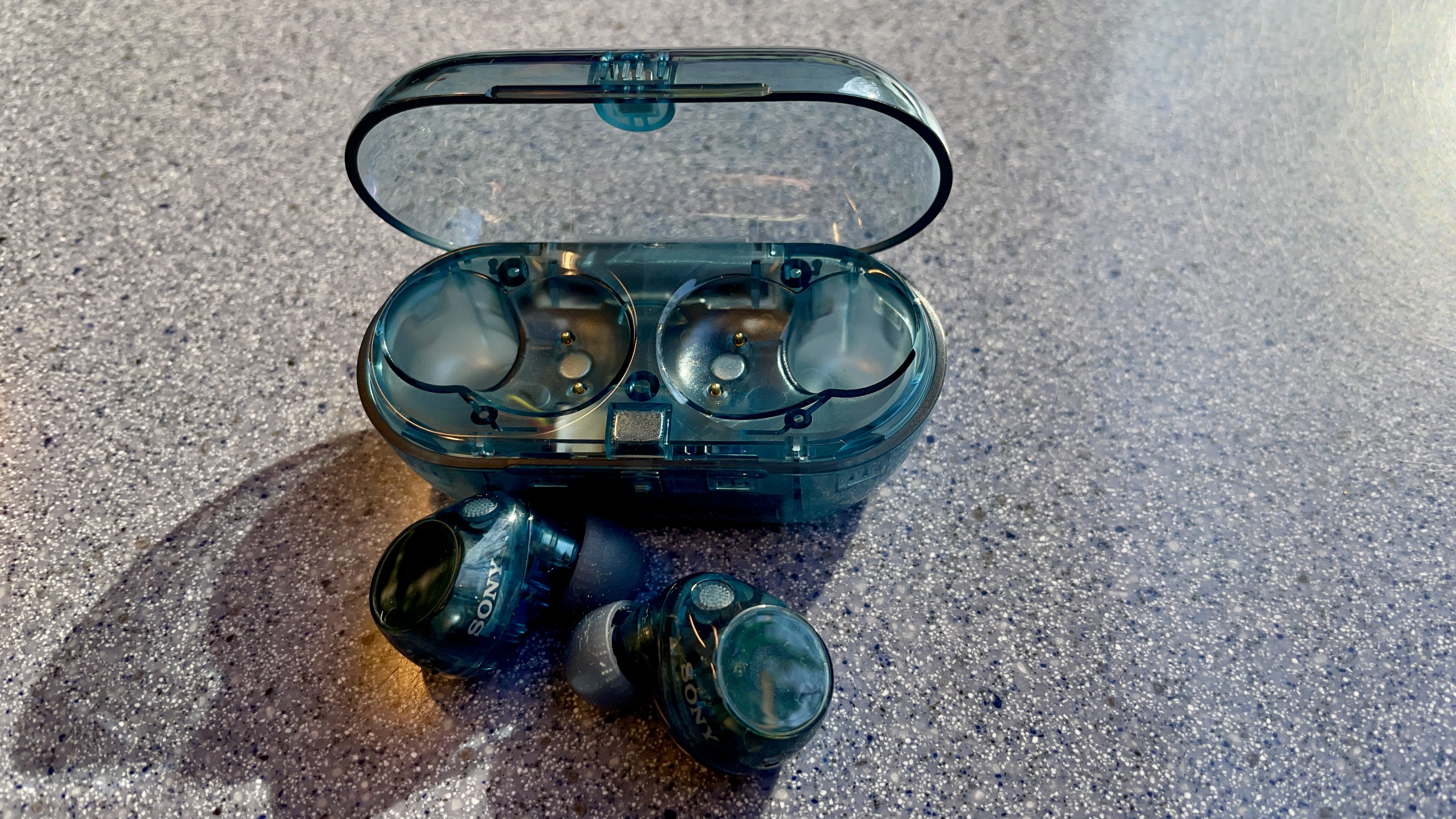 Sony WF-C710N earbuds in 'blue glass' on a grey table, with a woman's hand holding one of the earpieces